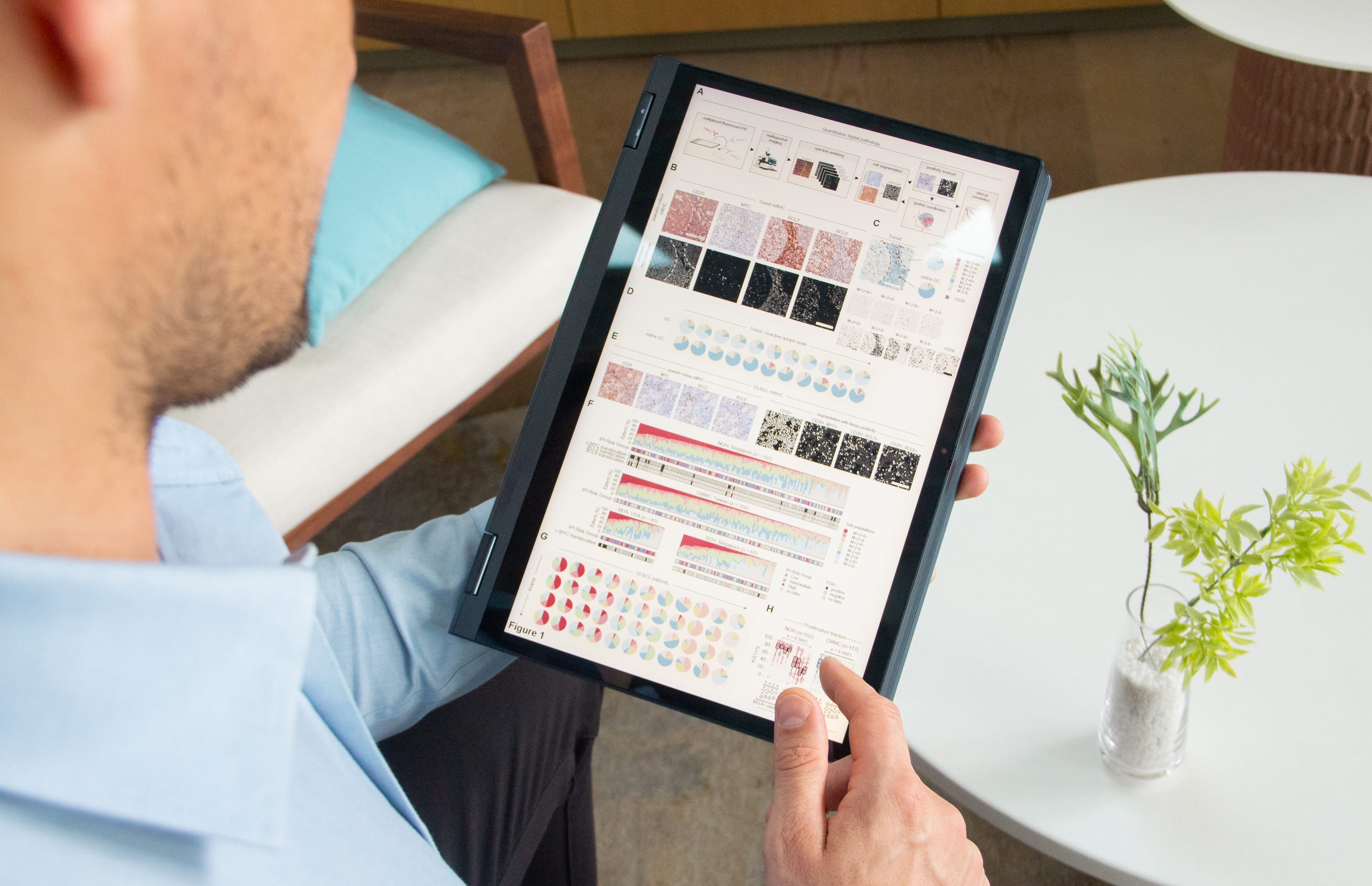 A person with facial hair holds a tablet displaying various scientific graphs and charts, sitting near a small plant.